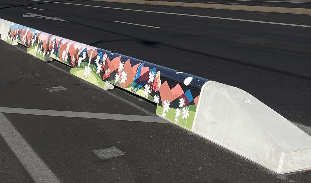 A concrete street barrier painted with a colorful mural of red mountains, green grass, and white flowers, located on an asphalt road with lane markings visible.