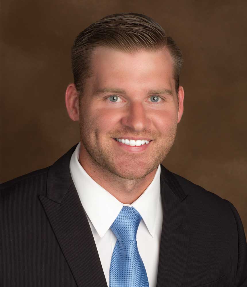 A man with short light brown hair, a trimmed beard, and blue eyes is smiling. He is wearing a black suit, white shirt, and light blue tie, posed against a brown background.