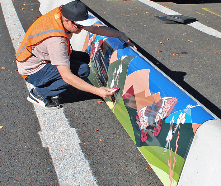 A person in an orange safety vest applies colorful butterfly and flower artwork to a concrete barrier on a sunny street.