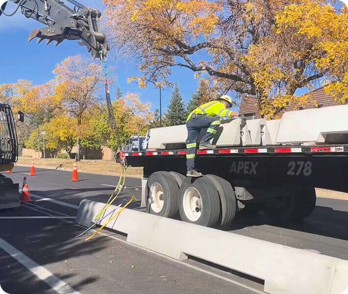 A construction worker in a high-visibility jacket steps onto a flatbed truck loaded with concrete barriers, while a crane lifts a barrier nearby. Trees with autumn leaves line the street. Orange cones mark the work area.