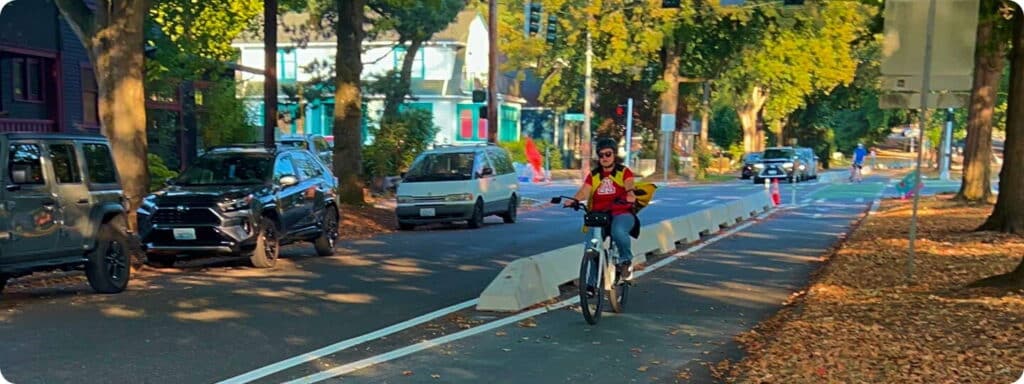 A person wearing a helmet and red shirt rides a bike in a protected bike lane on a tree-lined street, passing parked cars and colorful houses on a sunny day.