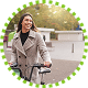 A woman wearing a gray coat smiles while standing outdoors with a bicycle. The image is framed by a green dotted border, and the background shows a blurred park or urban setting.