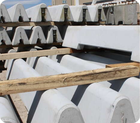 Stacks of large, white precast concrete beams and blocks are arranged outdoors, separated by wooden planks for support and stability. The scene suggests a construction or storage site.