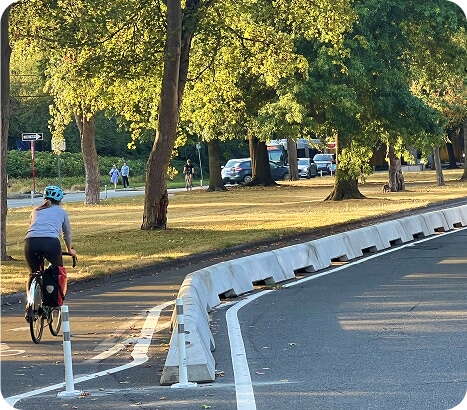 A person wearing a helmet rides a bicycle along a separated bike lane bordered by white barriers in a park-like area with green trees and people walking in the background.