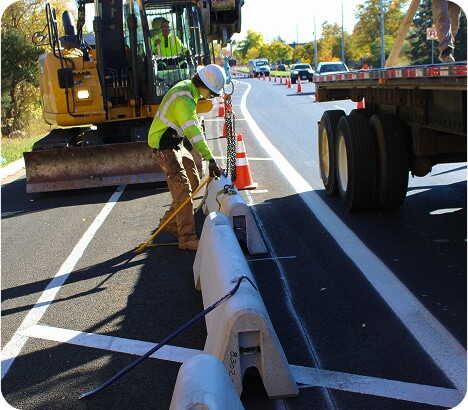 A construction worker in a safety vest and helmet adjusts concrete barriers along a road, with traffic cones, a truck, and construction equipment nearby.