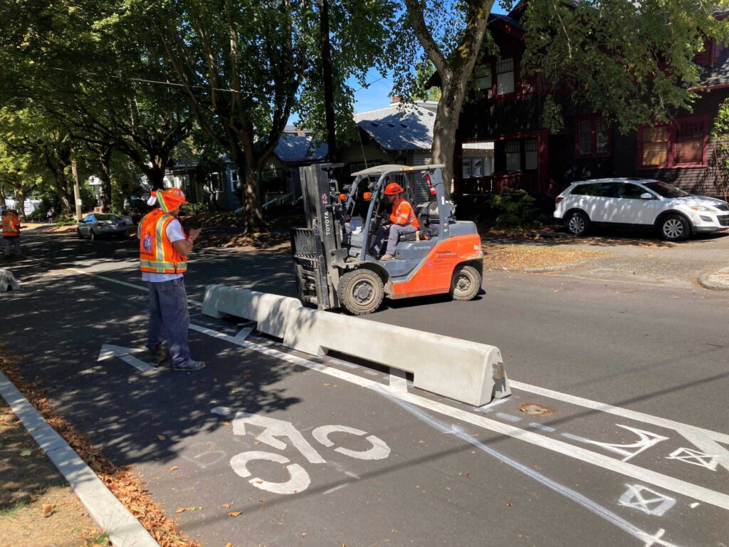 A construction worker guides a forklift operator as they place a concrete barrier along a bike lane on a tree-lined residential street.