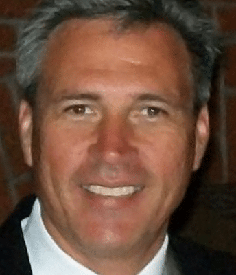 A middle-aged man with gray hair is smiling, wearing a dark suit jacket and a white shirt, standing in front of a brick wall background.