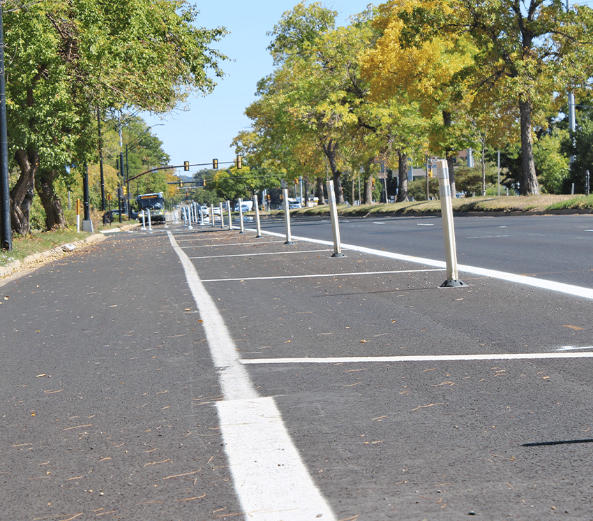 A freshly paved street with a separated bike lane marked by flexible white posts and white lines, bordered by trees with green and yellow leaves on a sunny day. Traffic signals are visible in the distance.