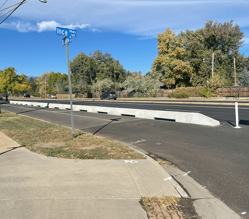 A street view shows a new concrete median separating traffic lanes on a sunny day, with a street sign reading "Inca Pkwy" and trees with green and yellow leaves in the background.