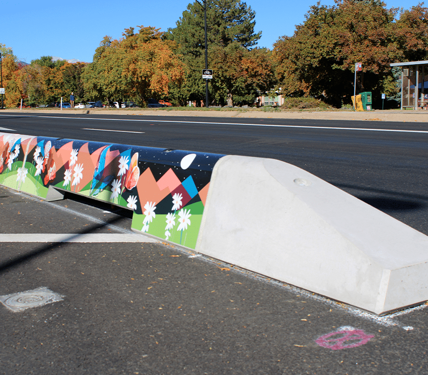 A concrete barrier by a road features colorful artwork of mountains, flowers, and trees under a partly sunny sky. There are autumn trees, a bus stop, and a clear blue sky in the background.