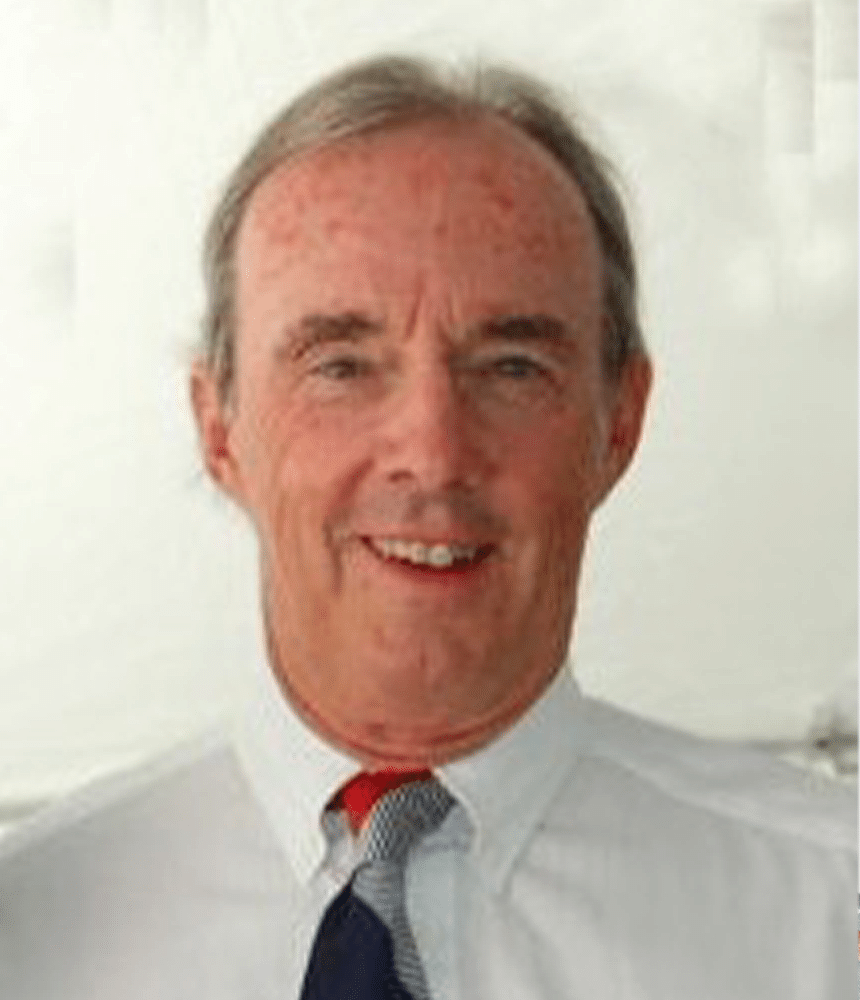 A middle-aged man with gray hair, wearing a white shirt and a red, white, and blue striped tie, smiles at the camera against a light background.