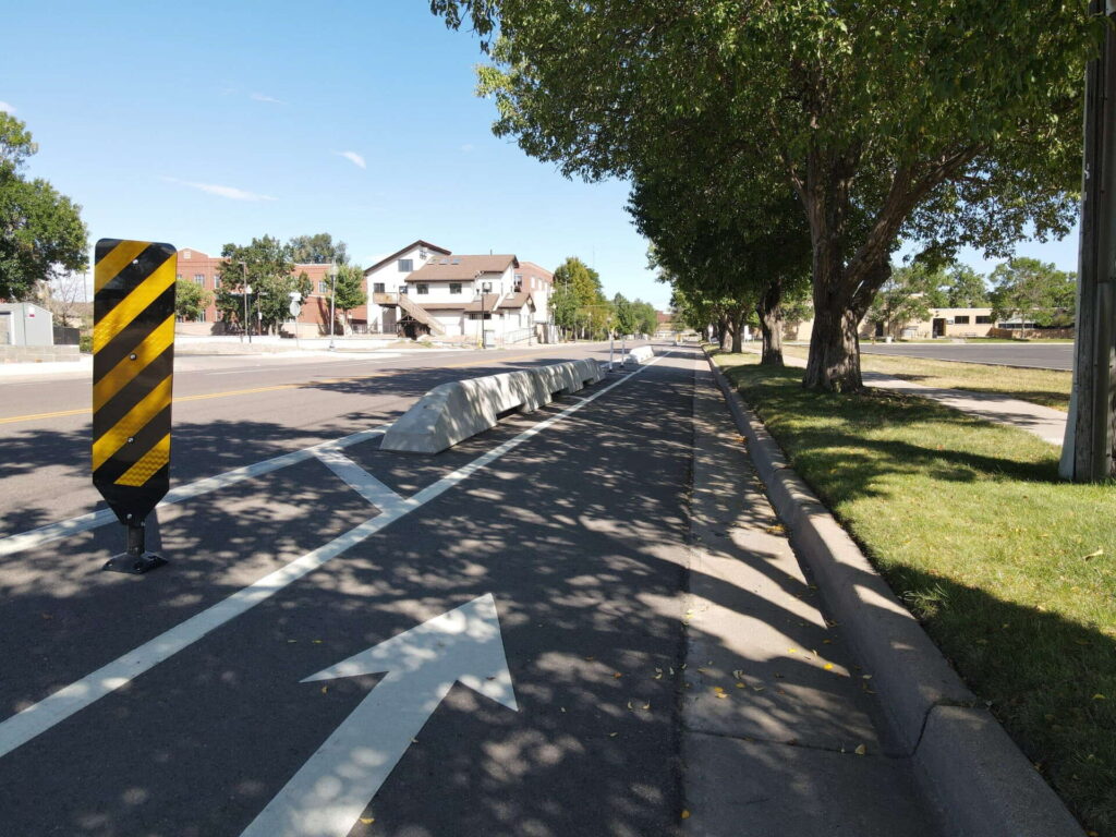 A separated bike lane runs alongside a street with white barriers and an arrow painted on the pavement. Trees line the sidewalk, and houses are visible in the background under a clear sky.