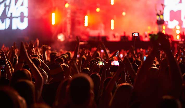 A crowd of people with raised hands enjoys a concert, with many holding up phones to record. Bright red and orange stage lights illuminate the scene in the background.