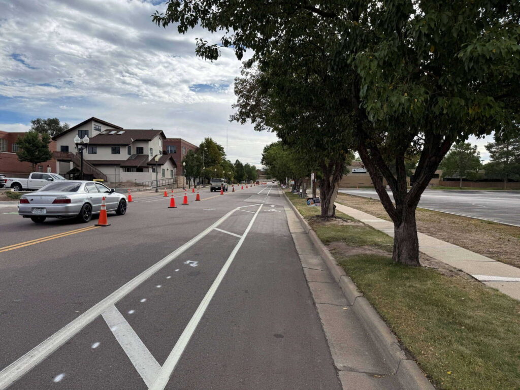 A street with a bike lane runs alongside parked cars and orange traffic cones, with trees lining the sidewalk and buildings visible in the background under a partly cloudy sky.
