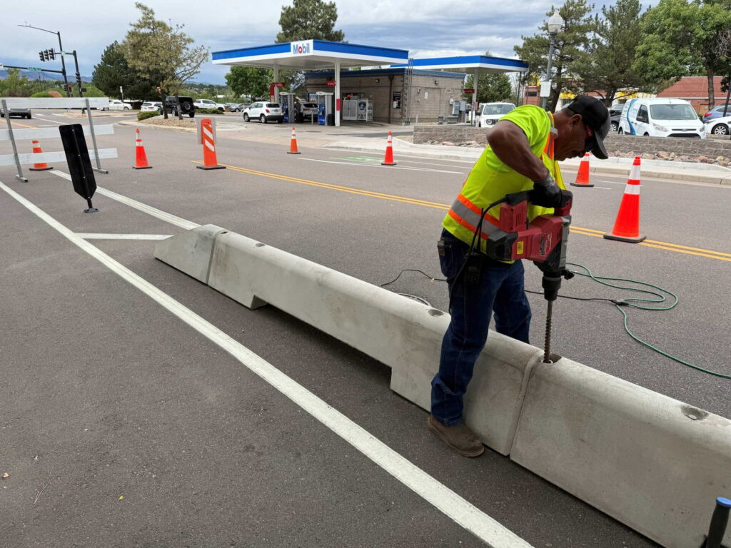 A construction worker in a yellow safety vest uses a power tool to drill into a concrete barrier on a street, with traffic cones and a gas station visible in the background.