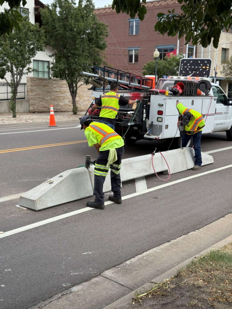 Three road workers in safety vests install concrete barriers along a street next to a parked utility truck with equipment. An orange traffic cone is placed nearby for safety. Trees and buildings are in the background.