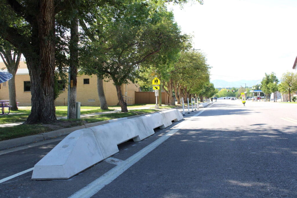 A row of large concrete barriers separates a bike lane from a street lined with trees and buildings on a sunny day. A pedestrian crossing sign is visible in the background.