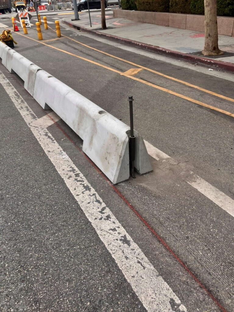 A concrete barrier separates a bike lane from the road. A long, thin metal bolt sticks upright from the barrier, posing a hazard. Yellow bollards, traffic cones, and a tree are visible in the background.