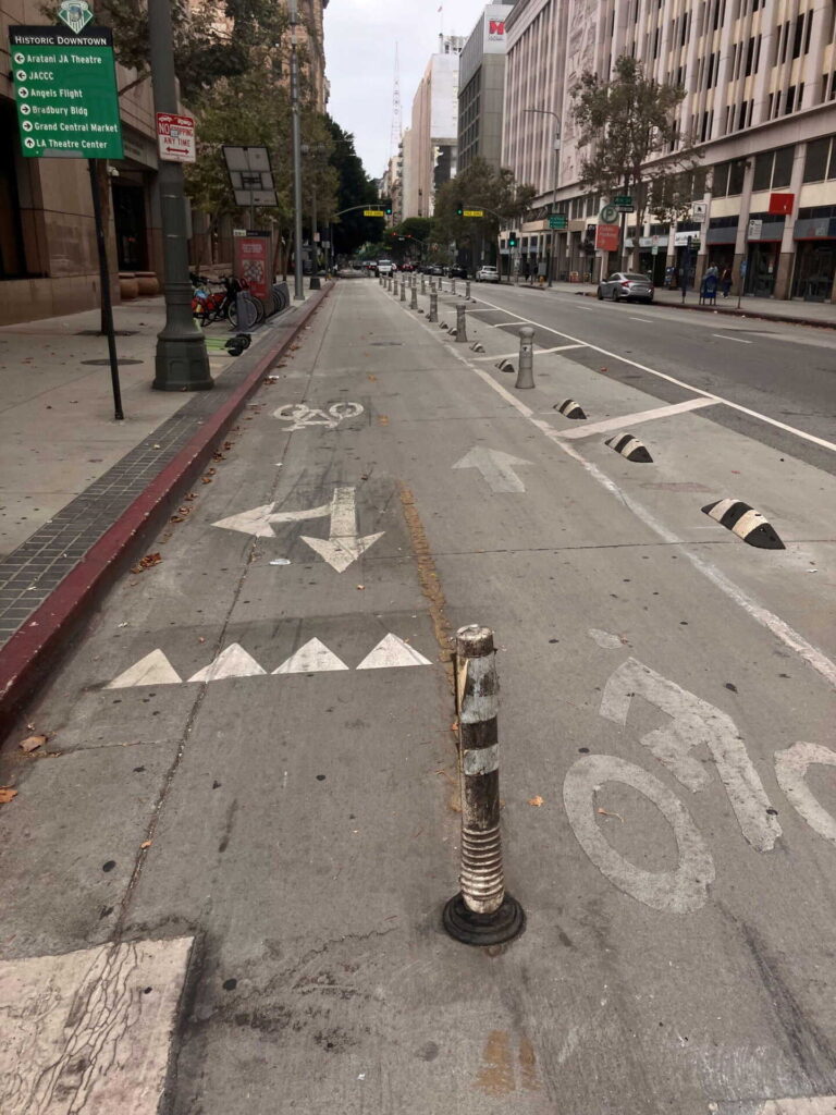 A wide bike lane on a city street is separated from traffic by flexible bollards and concrete barriers. Directional arrows and bike symbols are painted on the pavement. Tall buildings and trees line the empty street.