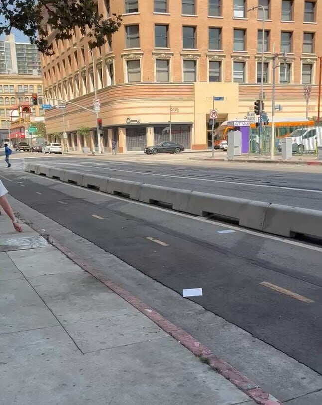 A city street with a bike lane separated by concrete barriers, a large corner building in the background, and a few scattered papers on the pavement. The street is empty and there are no vehicles in sight.
