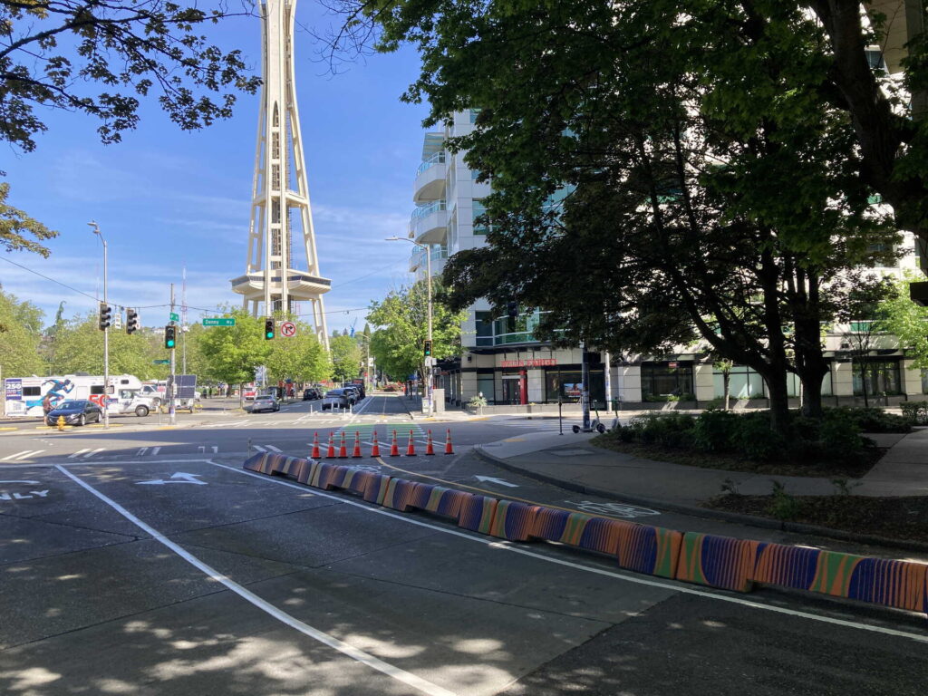 The Space Needle rises in the background near a city street lined with trees, traffic cones, and colorful road barriers. Cars and buildings are visible under a clear blue sky.