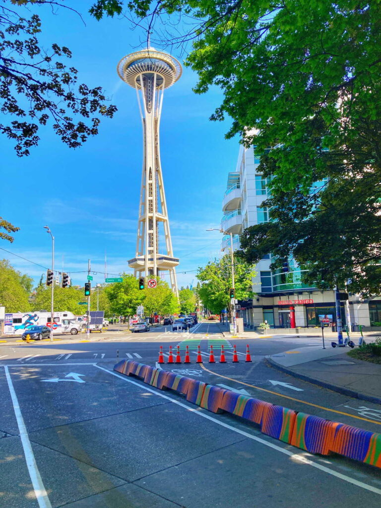 The Space Needle rises above a city street lined with trees, buildings, and traffic cones near a colorful barrier under a bright blue sky.