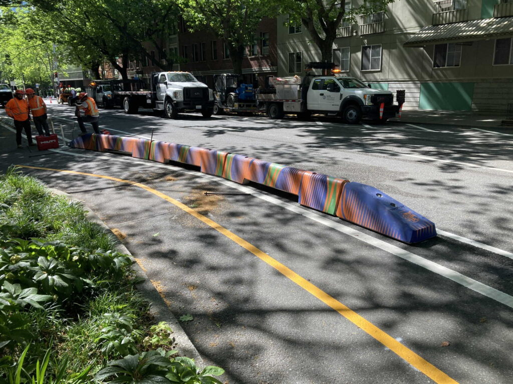 Workers install a colorful barrier separating a bike lane from the street. Trucks are parked nearby, and trees cast shadows on the road.