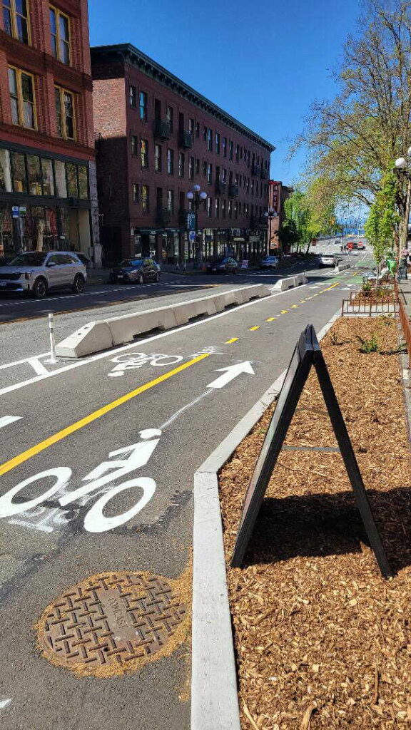 A city street with a separated bike lane marked by concrete barriers, bike lane symbols, and clear lane arrows. Cars are parked along the road, and buildings and trees line the sidewalks under a blue sky.