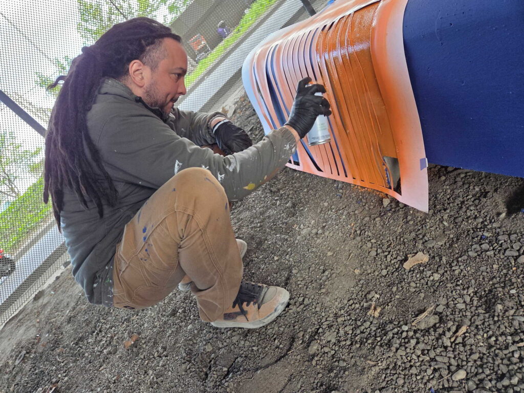 A person with dreadlocks crouches on gravel while spray painting an orange and blue design on a surface, using a stencil.