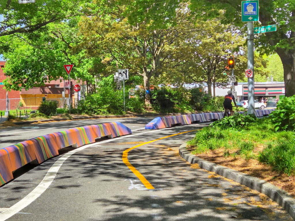 A curved bike lane with colorful barriers runs along a shady street lined with green trees. A cyclist waits at a red light, and several traffic signs are visible in the background.