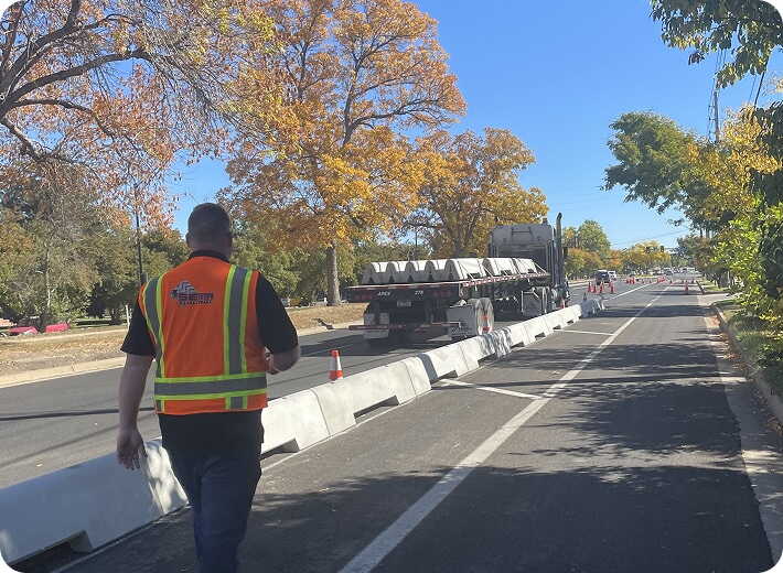 A person in a safety vest walks along a street with concrete barriers and construction equipment on a sunny day. Trees with autumn leaves line the road.