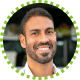 A man with short dark hair and a beard smiles at the camera. He is wearing a white shirt and is photographed outdoors with blurred greenery in the background. The image has a green dotted border.