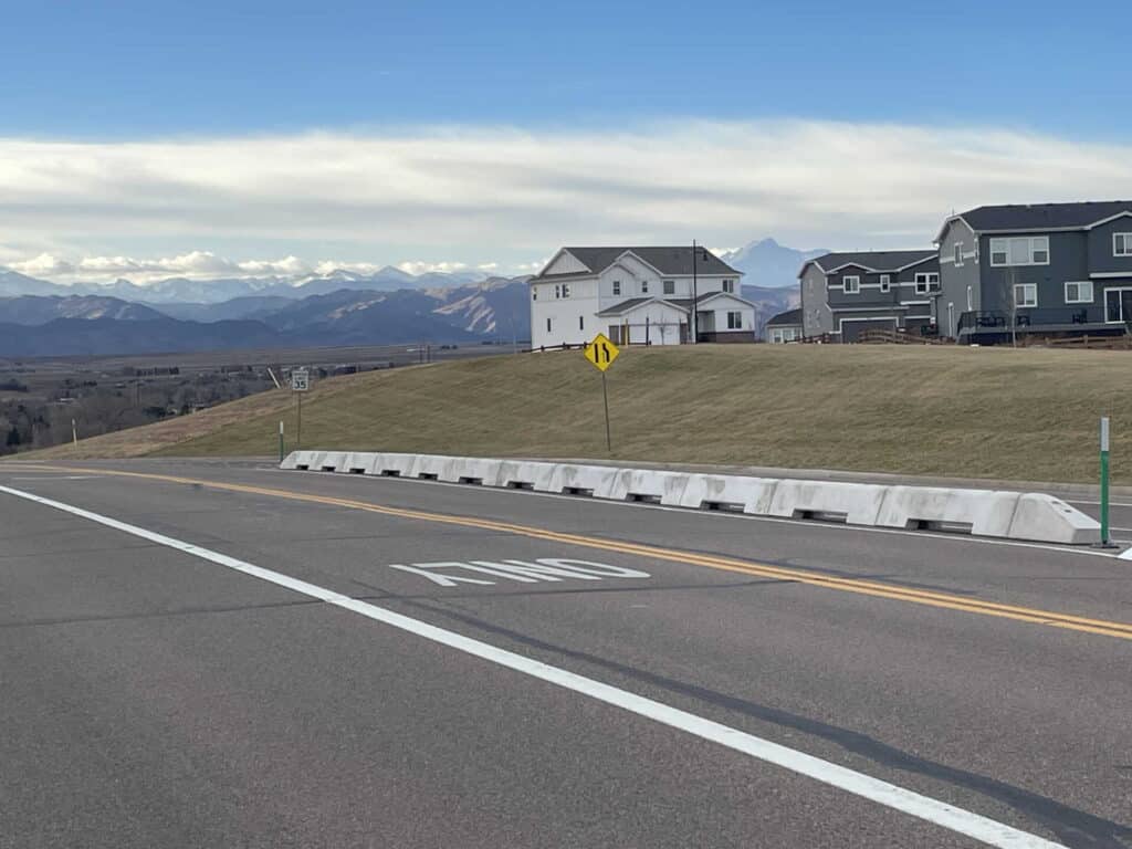 A paved road with a painted "END" marking curves left, bordered by concrete barriers. Suburban houses sit on a grassy hill, with mountains and a cloudy sky in the distance.