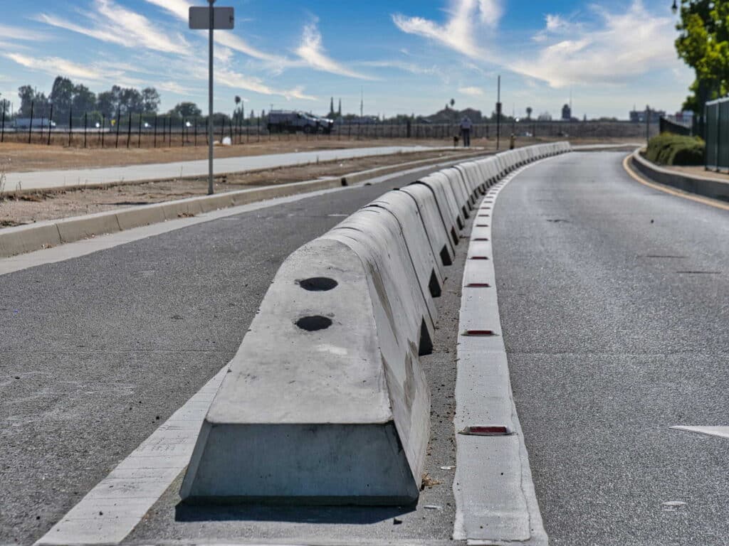 A row of concrete barriers separates two lanes on a curved road under a blue sky with wispy clouds; dry fields and a sidewalk are visible on the left.