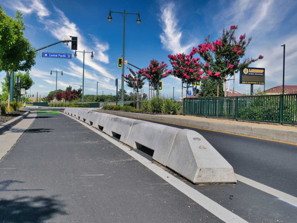 A protected bike lane divided from the main road by concrete barriers, with blooming pink trees, street lights, traffic signals, and street signs under a blue sky.