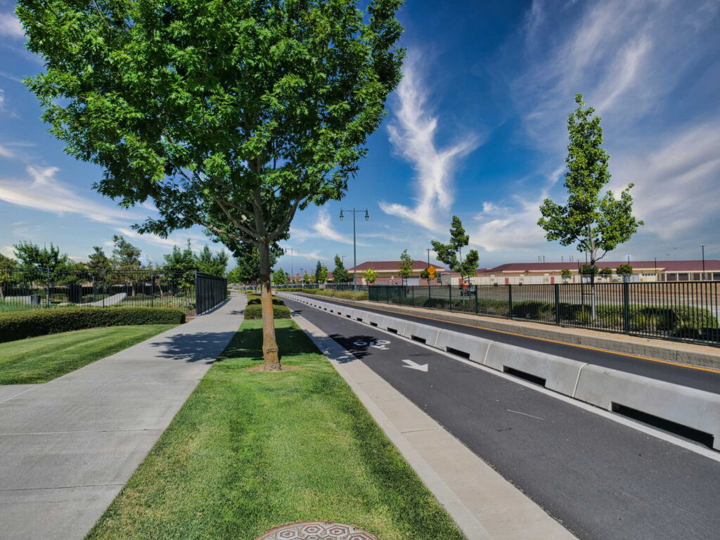 A tree-lined street with a bike lane, sidewalk, green grass, and a school or industrial building behind a black fence under a blue sky with wispy clouds.