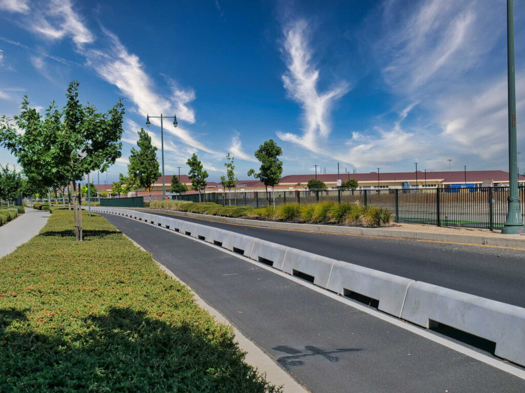 A tree-lined street with a bike lane, sidewalk, and barriers runs alongside a fence on a sunny day with blue sky and wispy clouds.