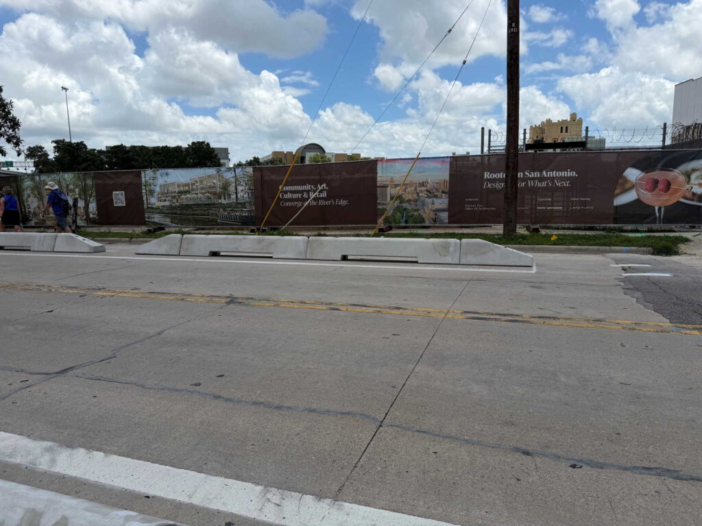 A street view of a construction site with brown fencing featuring promotional banners. The sky is partly cloudy, and a few people are walking on the sidewalk to the left. Concrete barriers separate the street from the site.