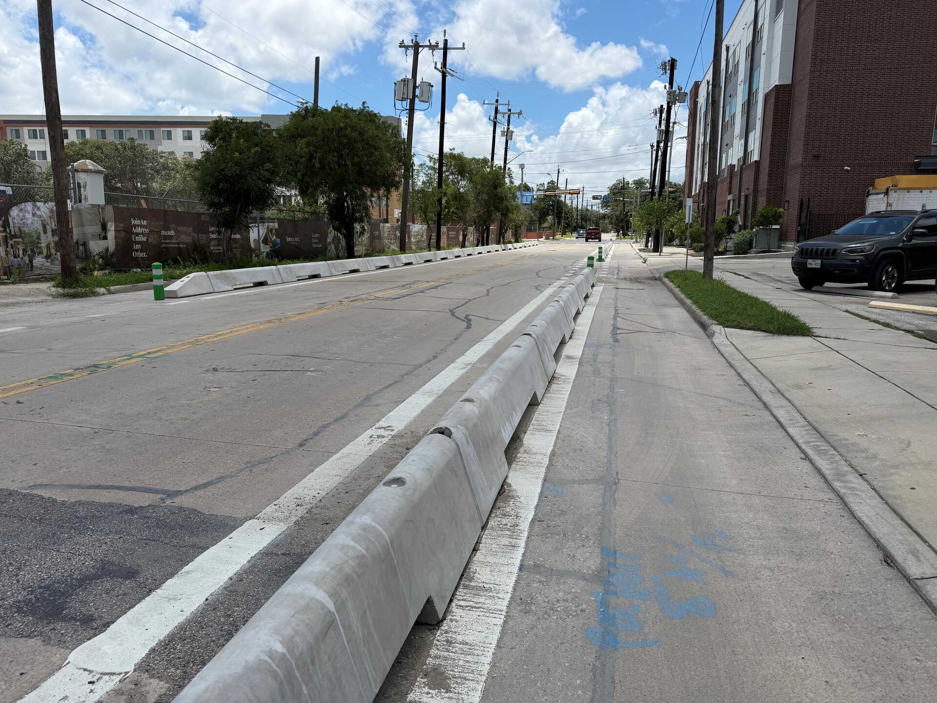 A city street with concrete barriers separates a bike lane from the road. Buildings, parked cars, and utility poles line the sidewalk under a partly cloudy sky.